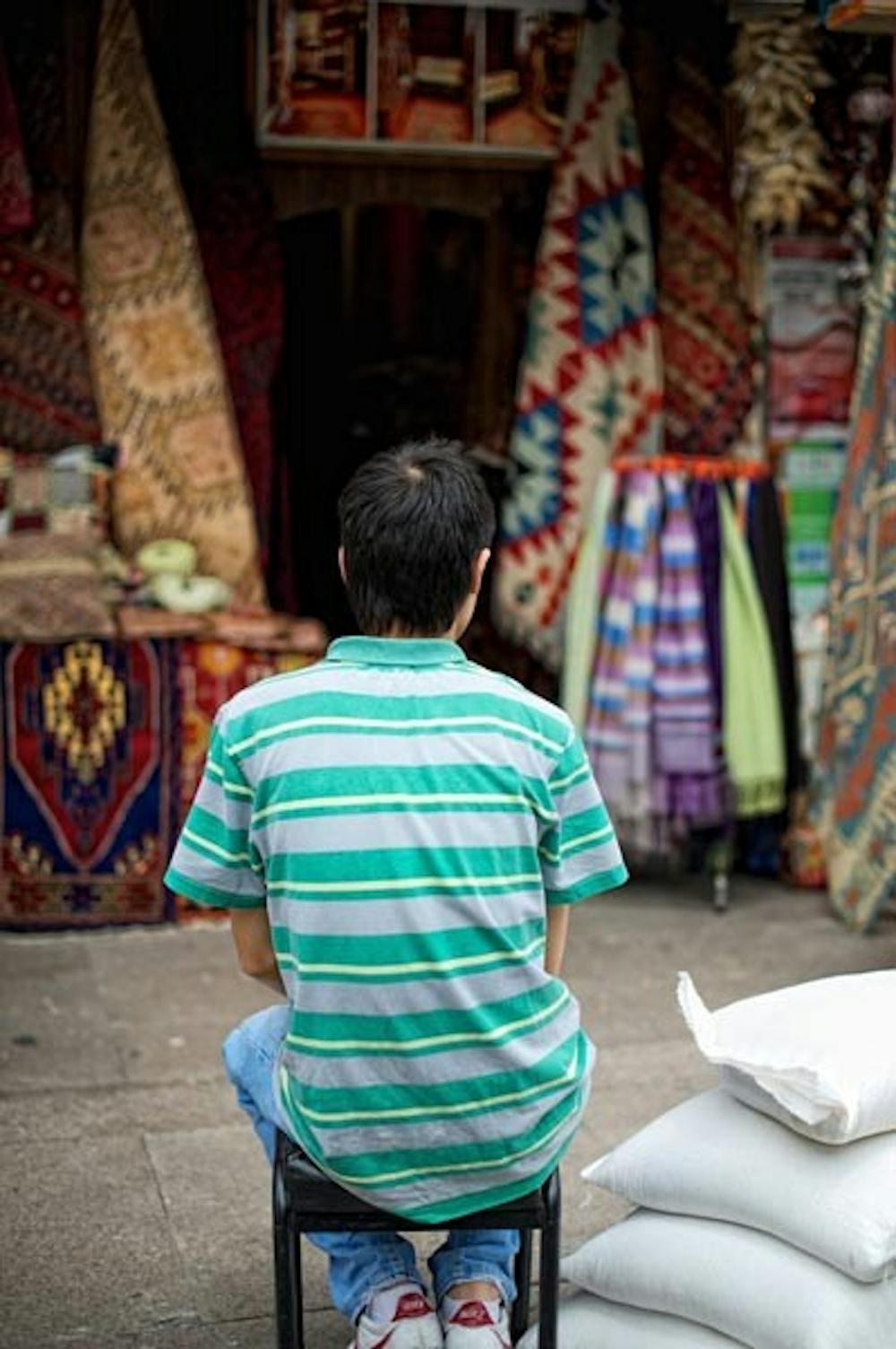 GLOBAL LOANS: A young boy waits for customers in front of his carpet stall at an outdoor bazaar in Selcuk, Turkey. Microloan organizations such as Kiva target similar individuals and small businesses to offer small-scale loans to. (Photo by Michael Arellano)