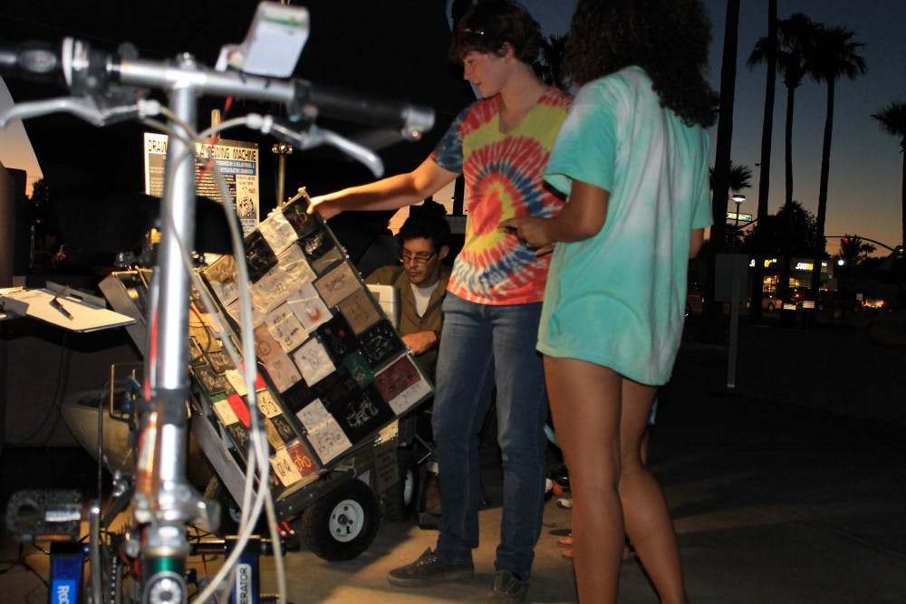ASU Art Museum visitors sift through Paul Nosa's collection of sewed works as he works on a personalized patch. The bicycle shown is used to generate power for his Singer sewing machine.  (Photo by Kylie Gumpert)