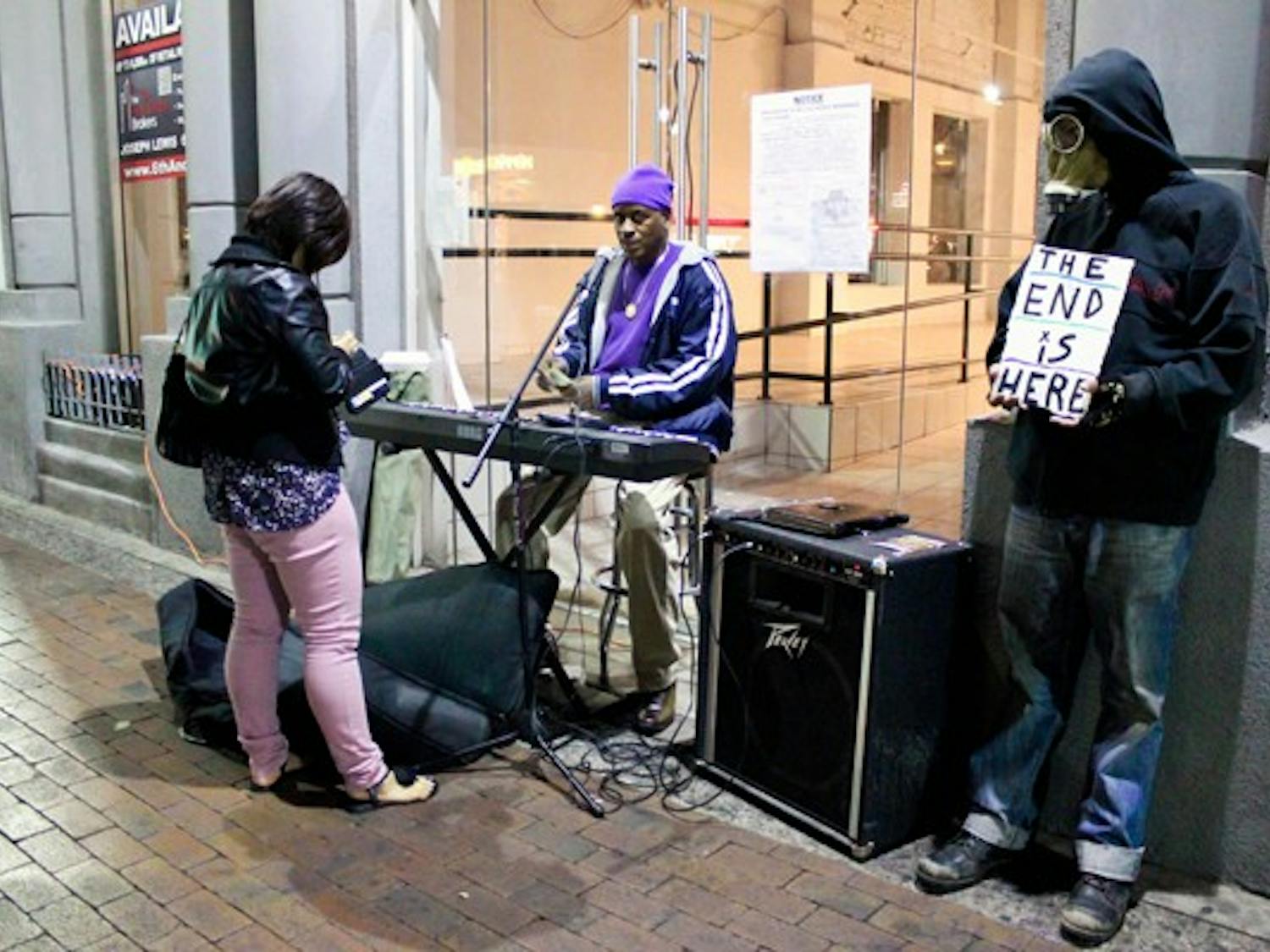 A man in purple, who refused to disclose his name, performs on Mill Avenue for extra cash while a homeless man named Sideshow wearing a gas mask holds a sign informing bystanders that “The End is Here.” (Photo by Marissa Krings)