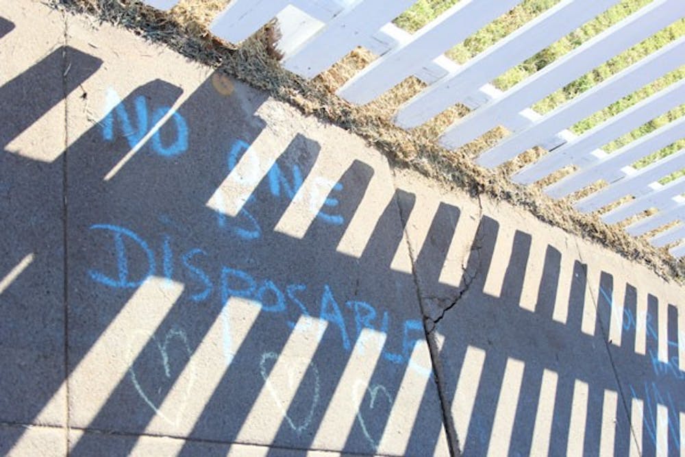 Shadows of a newly painted, white picket fence cover the chalk written words "No One is Disposable" on a sidewalk in Phoenix. (Photo by Ana Ramirez)