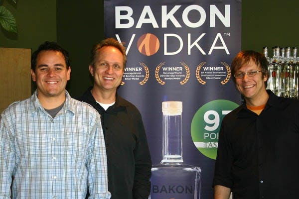 BAKON ON THE ROCKS: The creators of Bakon Vodka, Stefan Schachtell, Steve Liden and Chris Marshall (from left to right), pose in front of a banner endorsing their smoky beverage. Liden, an ASU electrical engineering graduate, co-founded the bacon-flavored vodka company that is currently available in 30 states and is soon arriving in Arizona.  (Photo courtesy of Bakon Vodka)