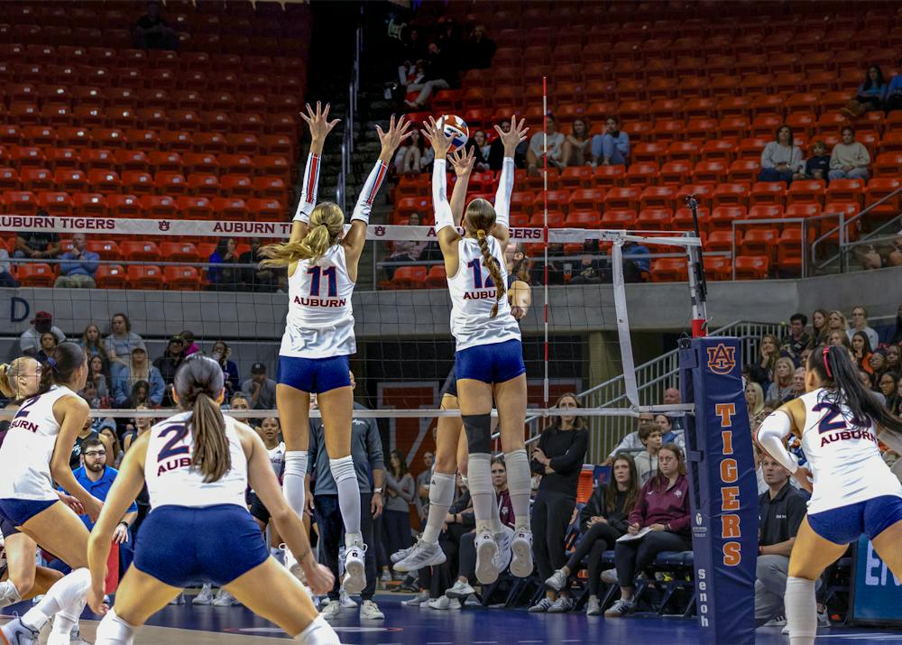 Auburn volleyball players in white jerseys and blue shorts jump at the net to block a volleyball during a match. Teammates prepare behind them as fans watch from the stands.