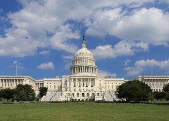 The U.S. Capitol building stands beyond a broad green lawn beneath a blue, partly cloudy sky.