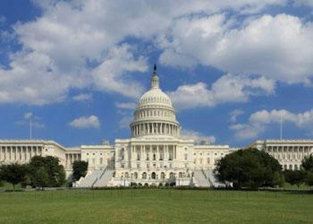 The U.S. Capitol building stands beyond a broad green lawn beneath a blue, partly cloudy sky.