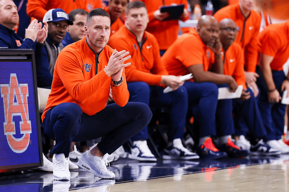 Auburn men’s basketball coach crouches near the sideline with hands clasped, focused on the court, while assistant coaches in orange team gear sit behind him on the bench during a game.