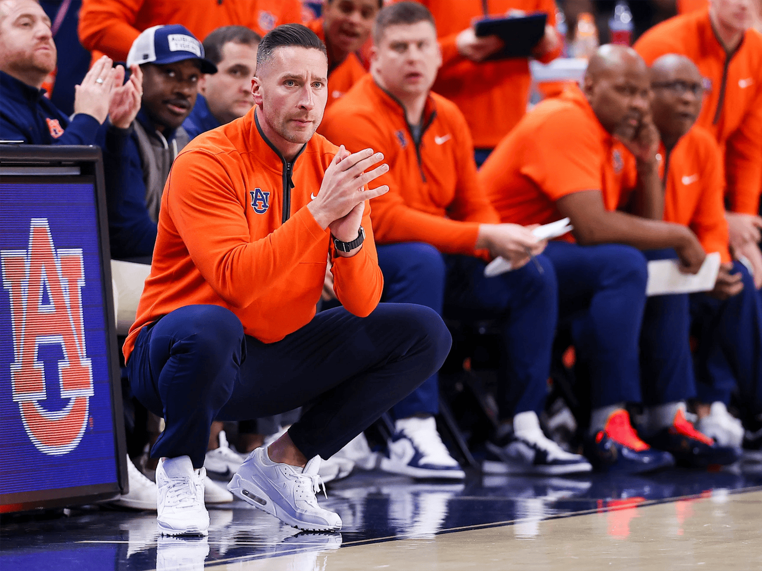 Auburn men’s basketball coach crouches near the sideline with hands clasped, focused on the court, while assistant coaches in orange team gear sit behind him on the bench during a game.