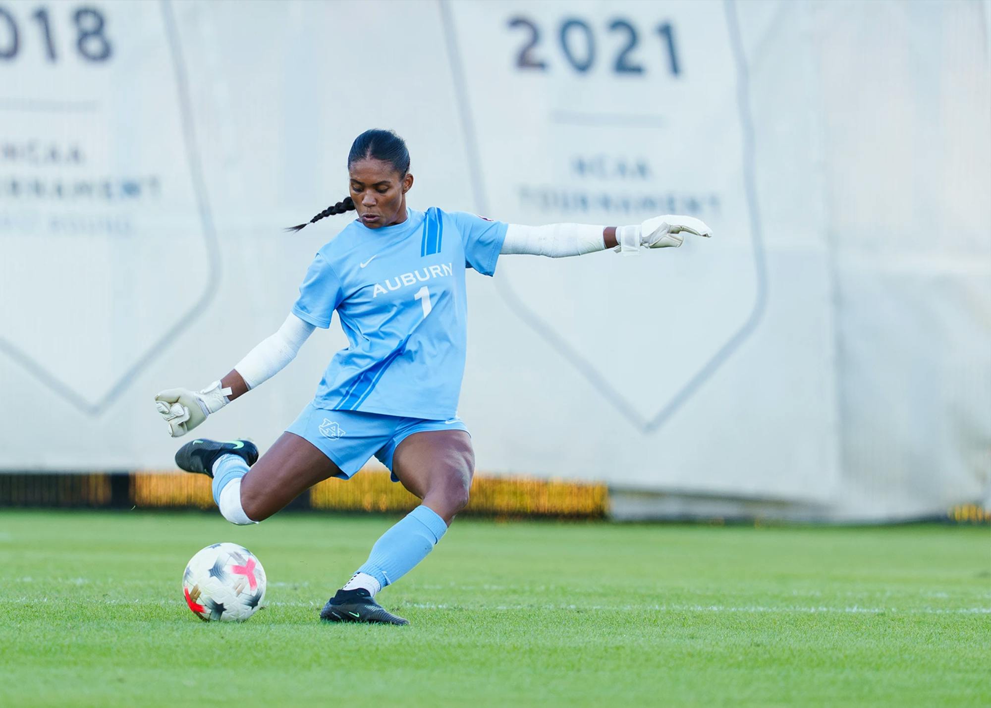 Auburn soccer goalkeeper Ayana Yapo mid-action on a green field, wearing a light blue kit with "AUBURN" and the number 1 printed on the chest. She has her hair styled in a braid and is wearing white goalie gloves and arm sleeves. Her right leg is extended mid-kick as she strikes a white soccer ball with a red and black pattern. Banners reading "2018" and "2021 NCAA TOURNAMENT" are visible in the background.