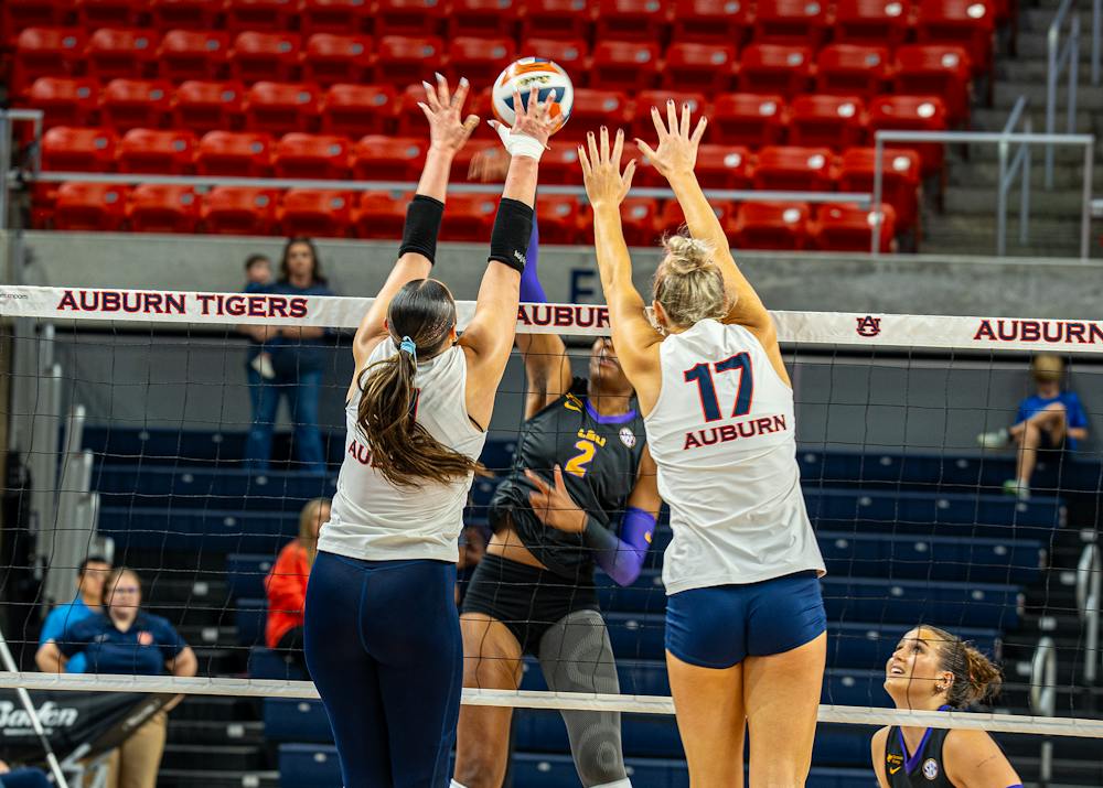 Auburn volleyball players Grace Havlicek (left) and Belle Patrick (right, wearing jersey No. 17) jump together at the net, raising their arms high to attempt a double block against LSU hitter Jurnee Robinson (No. 2) in a dark uniform. The action takes place during a match inside Neville Arena, with red arena seats in the background.