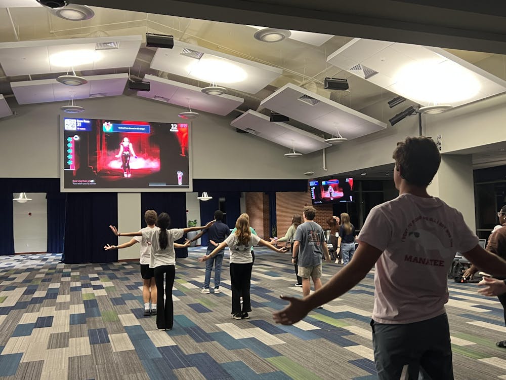 Group of students dancing in a large ballroom. They mimic the movements of a woman dancing on a large screen