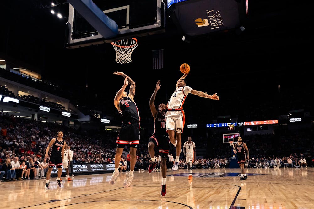 An Auburn basketball player leaps toward the basket with the ball extended in one hand during a fast-break play, while two defenders in black uniforms attempt to contest the shot. The action takes place on a brightly lit court inside a packed arena, with spectators watching from the stands and the scoreboard visible in the background.