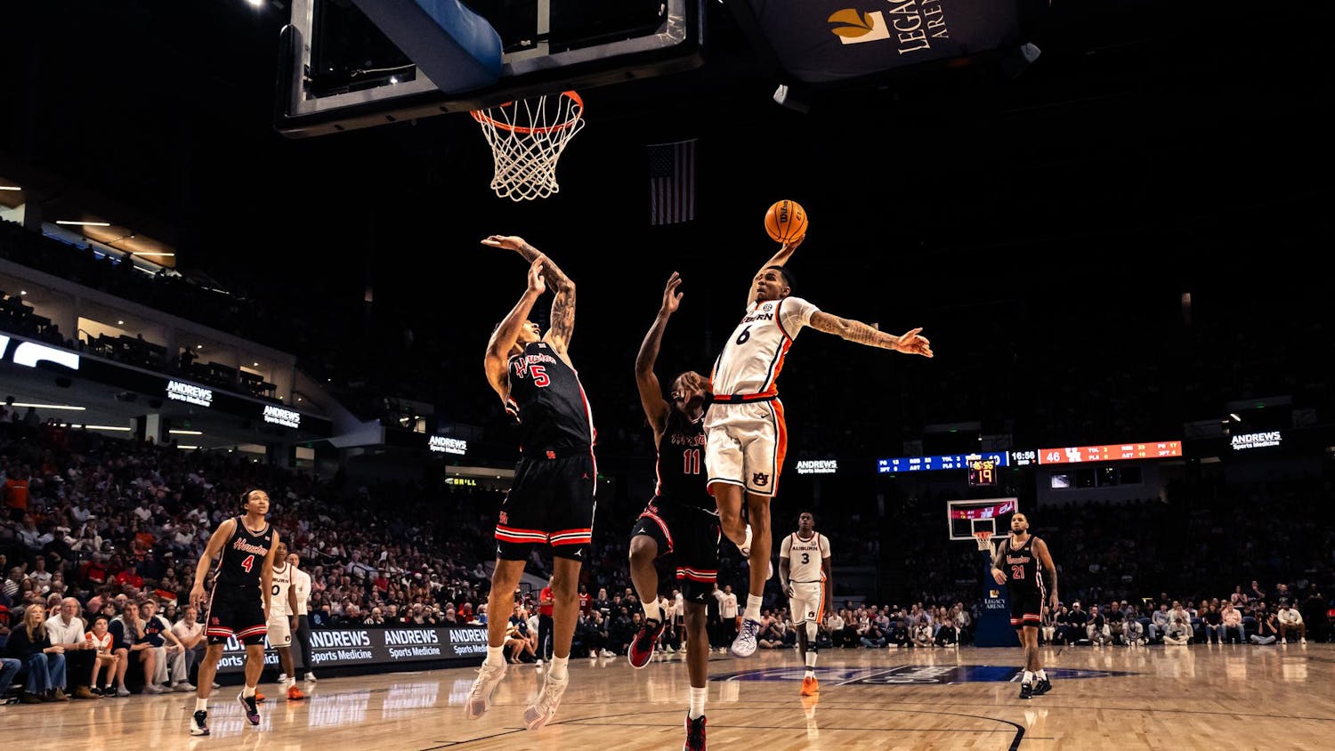 An Auburn basketball player leaps toward the basket with the ball extended in one hand during a fast-break play, while two defenders in black uniforms attempt to contest the shot. The action takes place on a brightly lit court inside a packed arena, with spectators watching from the stands and the scoreboard visible in the background.