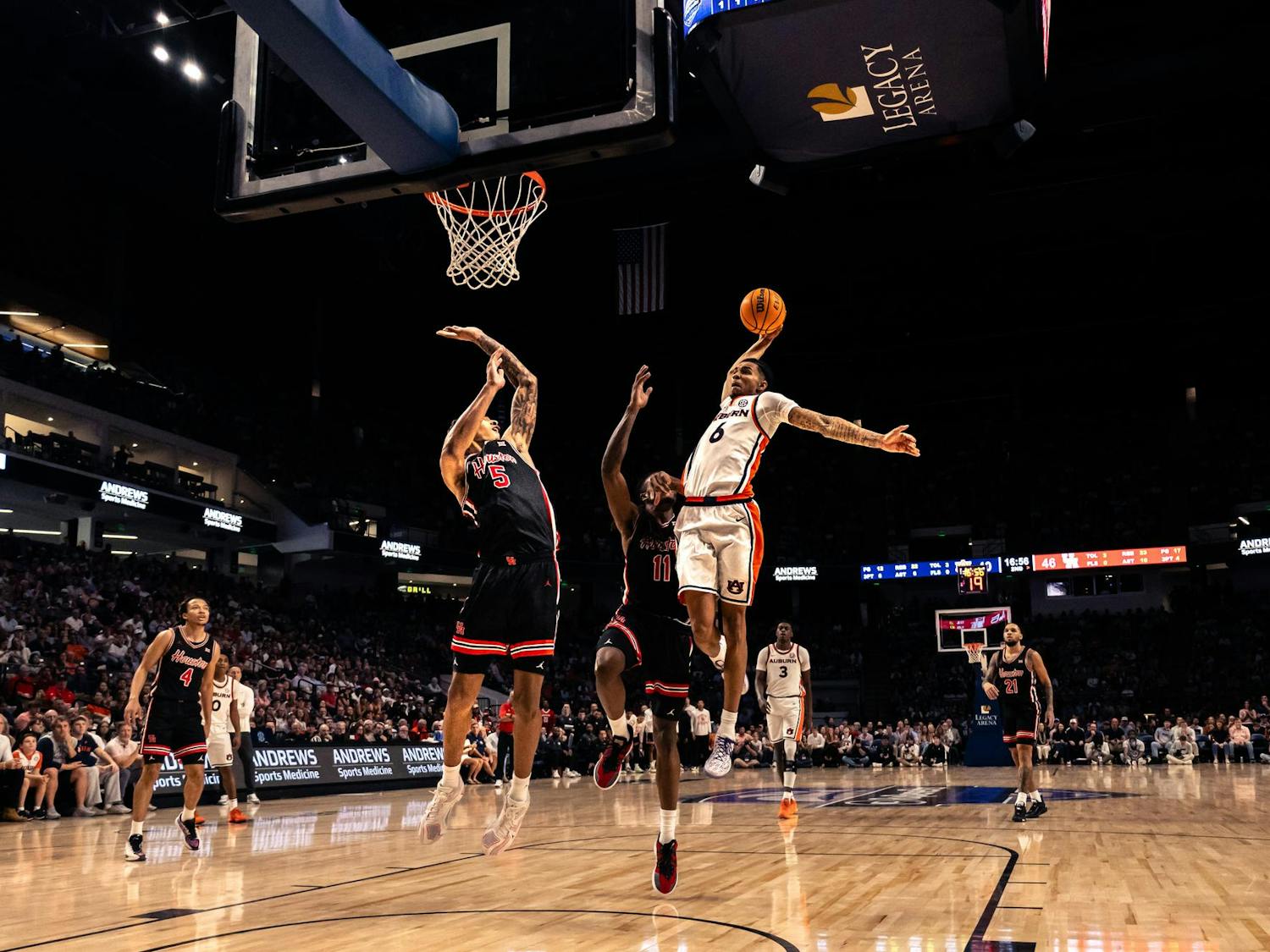 An Auburn basketball player leaps toward the basket with the ball extended in one hand during a fast-break play, while two defenders in black uniforms attempt to contest the shot. The action takes place on a brightly lit court inside a packed arena, with spectators watching from the stands and the scoreboard visible in the background.