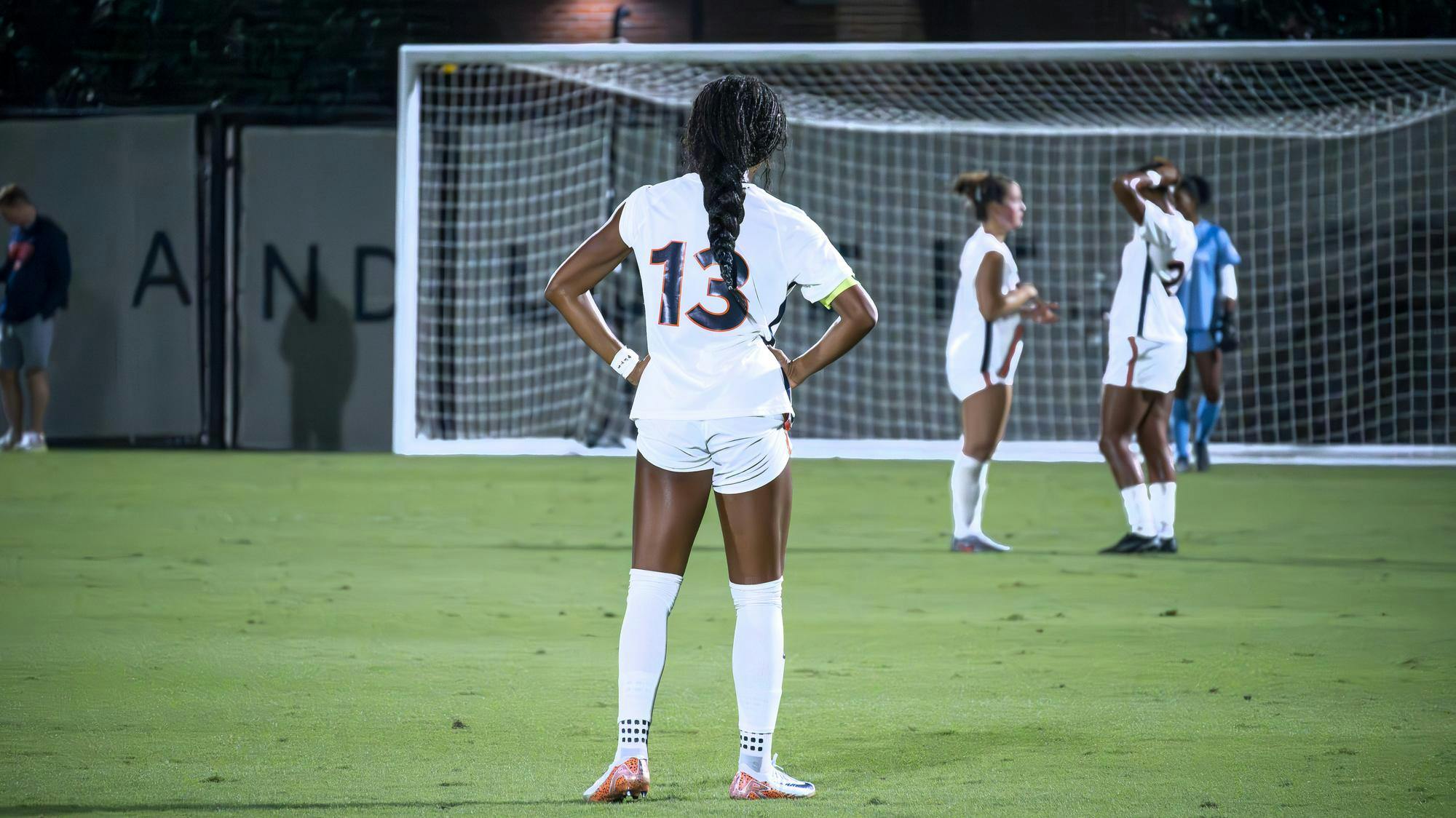 An Auburn soccer player wearing a white uniform with the number 13 stands with hands on hips facing the goal, while teammates in the background gather near the net during a nighttime match.