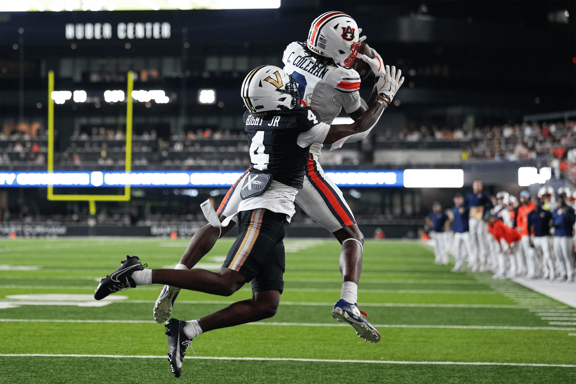 An Auburn football player wearing a white uniform leaps to catch a pass near the sideline while being closely defended by a Vanderbilt player in a black uniform. Both athletes are airborne as they battle for the ball, with the field, end zone, and bright stadium lights visible in the background.