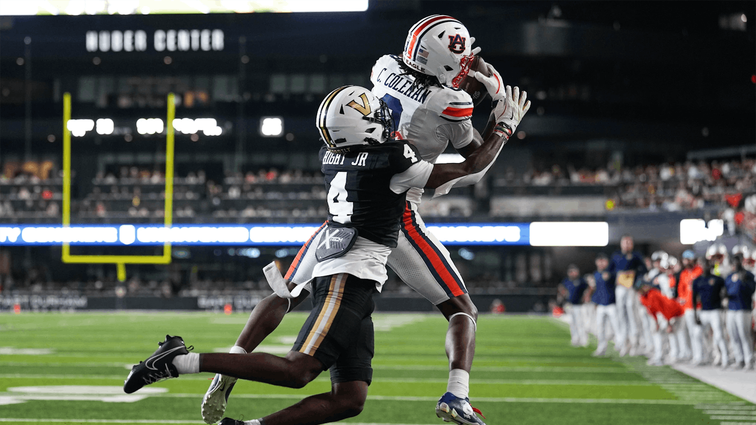 An Auburn football player wearing a white uniform leaps to catch a pass near the sideline while being closely defended by a Vanderbilt player in a black uniform. Both athletes are airborne as they battle for the ball, with the field, end zone, and bright stadium lights visible in the background.