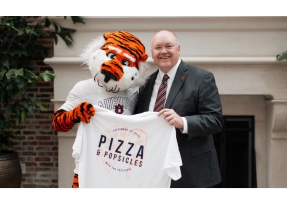 President Roberts stands next to Aubie as they both hold a t-shirt that says Pizza and Popsicles on top of slices of pizza and popsicles