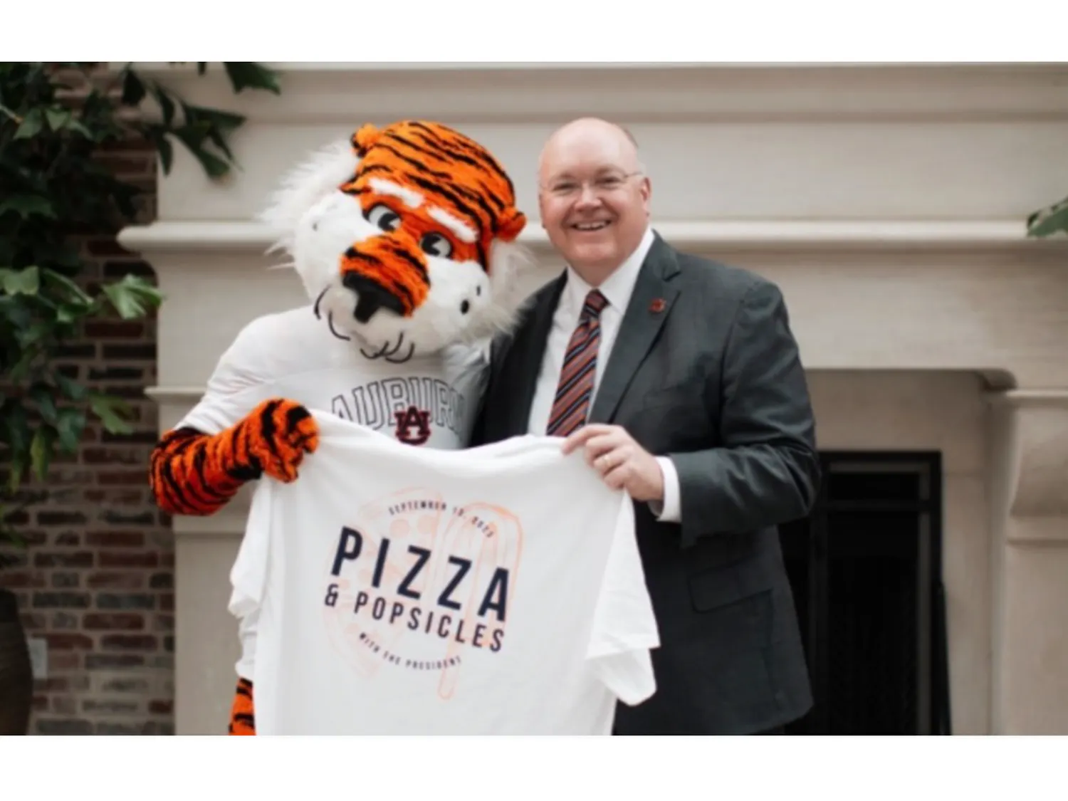 President Roberts stands next to Aubie as they both hold a t-shirt that says Pizza and Popsicles on top of slices of pizza and popsicles
