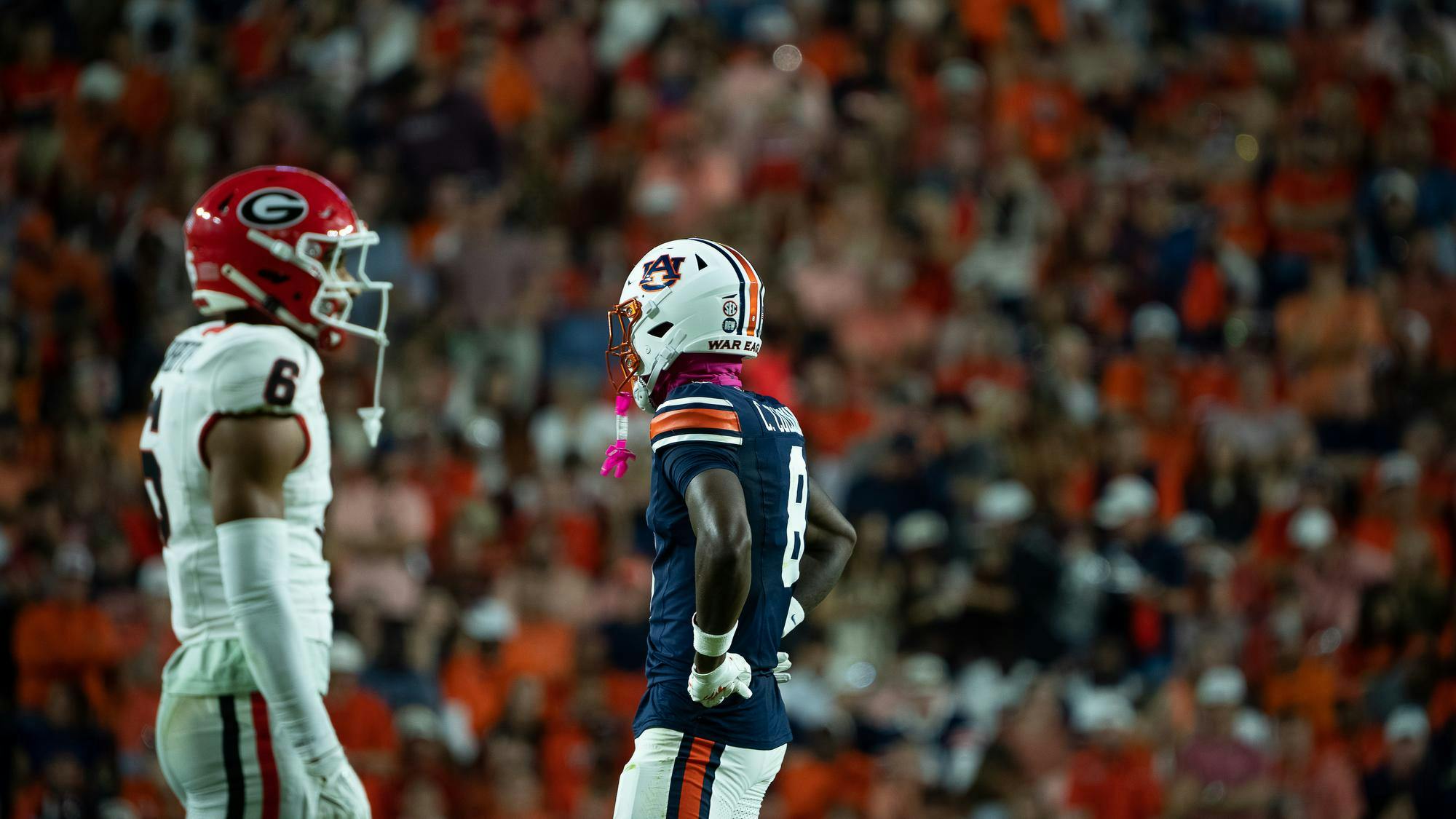 Auburn football player wearing a navy blue uniform and white helmet stands facing a Georgia player in a white uniform and red helmet during a game, with a blurred crowd in the background.