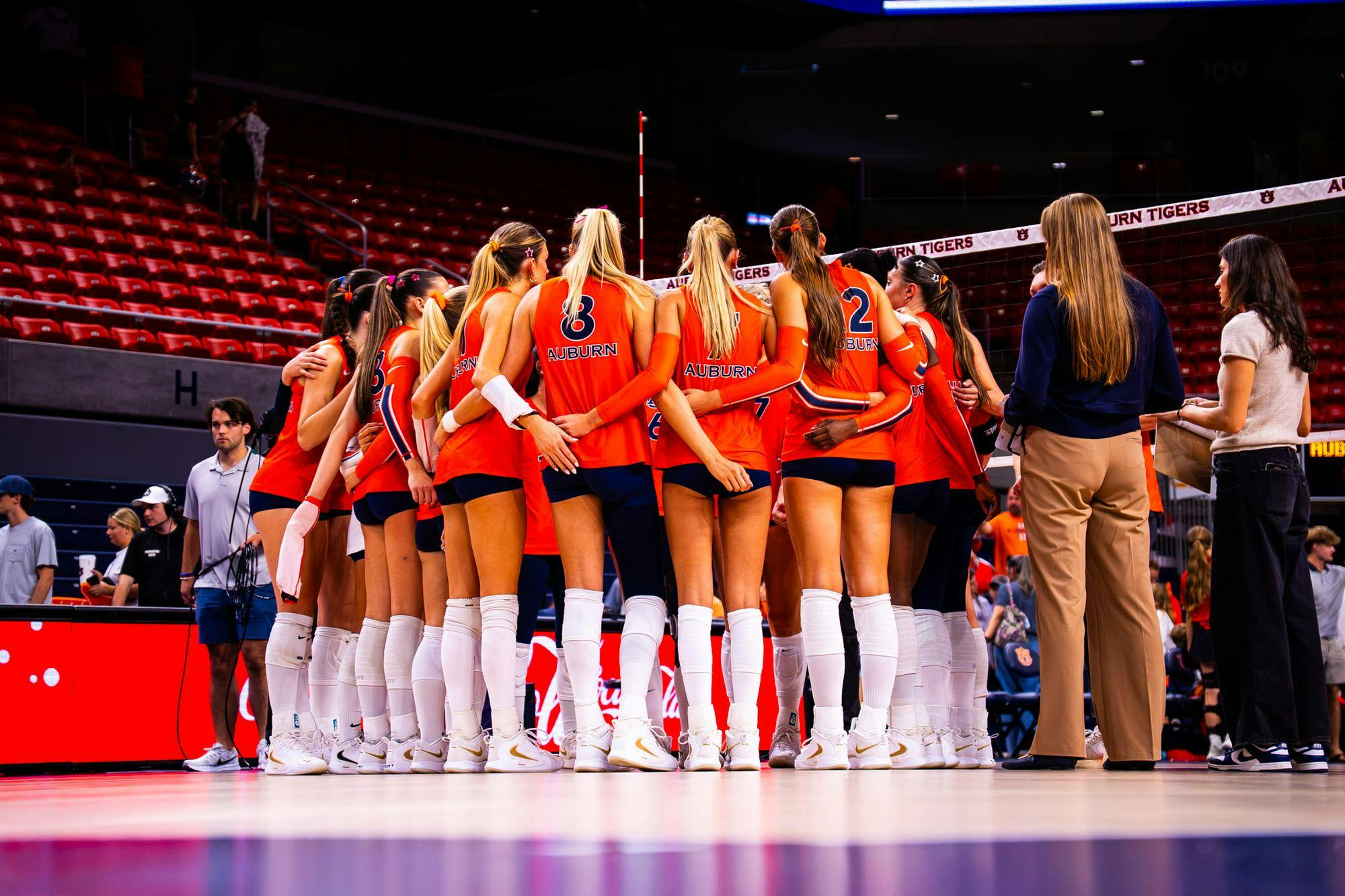 Auburn volleyball players huddle together with their arms around one another before a match, wearing orange jerseys, navy shorts, and white knee pads on the court.