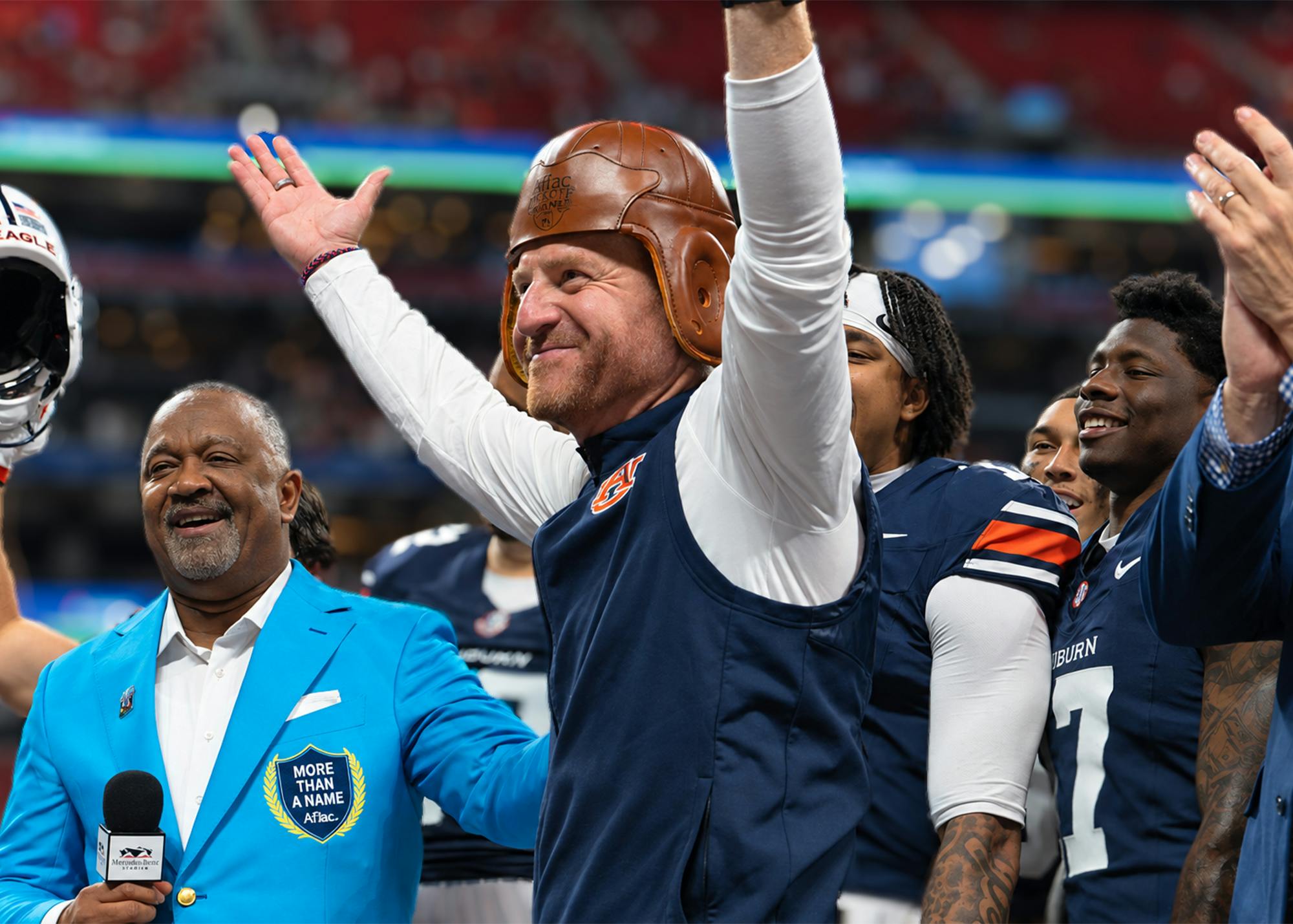 Alex Golesh celebrates Auburn’s victory at the Aflac Kickoff Game, wearing the Old Leather Helmet trophy on his head and raising his arms alongside Auburn players and Aflac representatives.