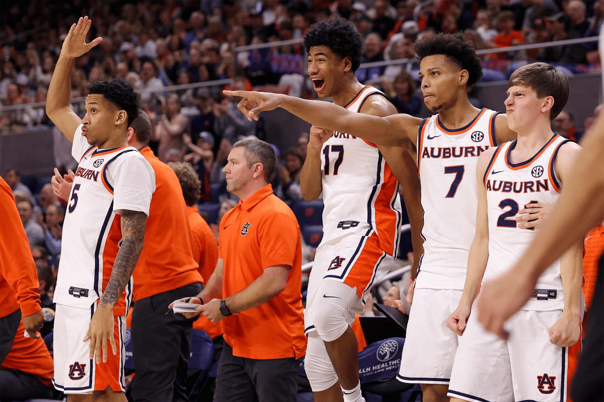 Auburn men’s basketball players in white uniforms stand and react excitedly on the sideline, with one player pointing toward the court and others cheering, while coaches in orange polos and a packed crowd fill the background.