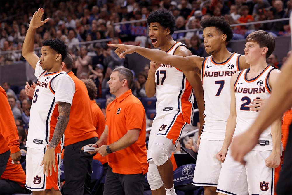 Auburn men’s basketball players in white uniforms stand and react excitedly on the sideline, with one player pointing toward the court and others cheering, while coaches in orange polos and a packed crowd fill the background.
