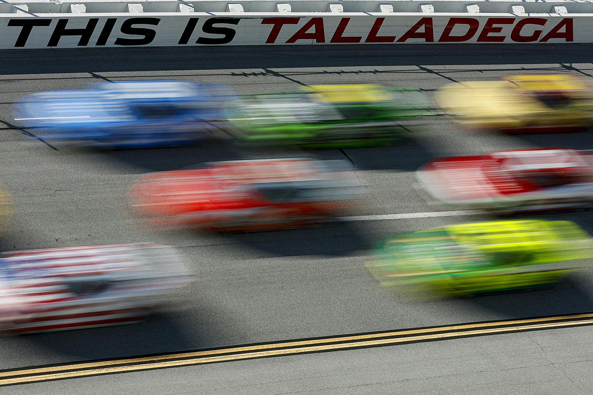 Blurred race cars in various bright colors speed down the track at Talladega Superspeedway, with the words “THIS IS TALLADEGA” visible on the wall in the background.