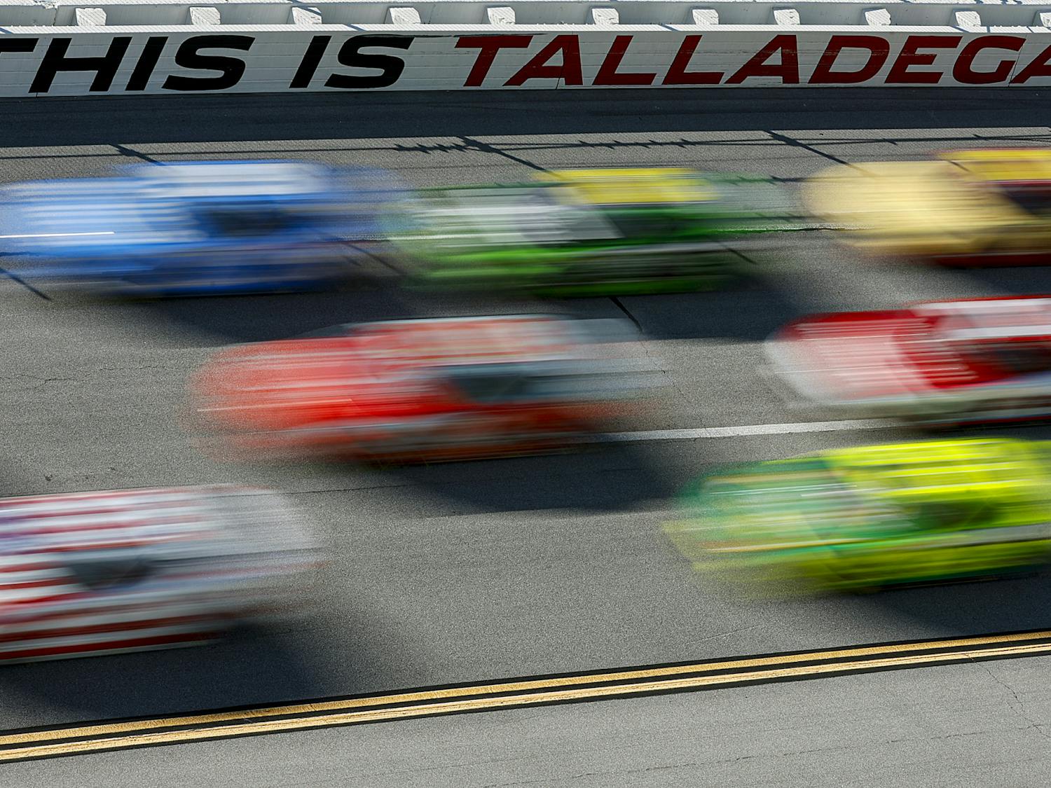 Blurred race cars in various bright colors speed down the track at Talladega Superspeedway, with the words “THIS IS TALLADEGA” visible on the wall in the background.