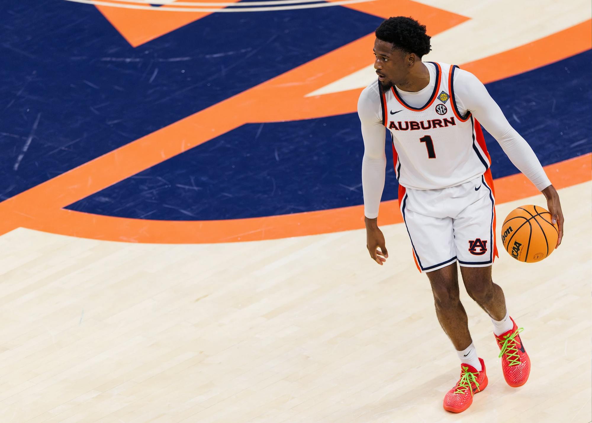 A male Auburn basketball player wearing a white jersey with the number 1 dribbles a ball on a hardwood court, looking to his left. He stands near a large orange and blue Auburn logo, wearing bright orange shoes with neon green laces.