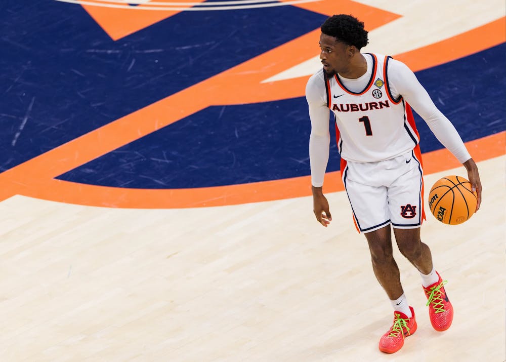 A male Auburn basketball player wearing a white jersey with the number 1 dribbles a ball on a hardwood court, looking to his left. He stands near a large orange and blue Auburn logo, wearing bright orange shoes with neon green laces.