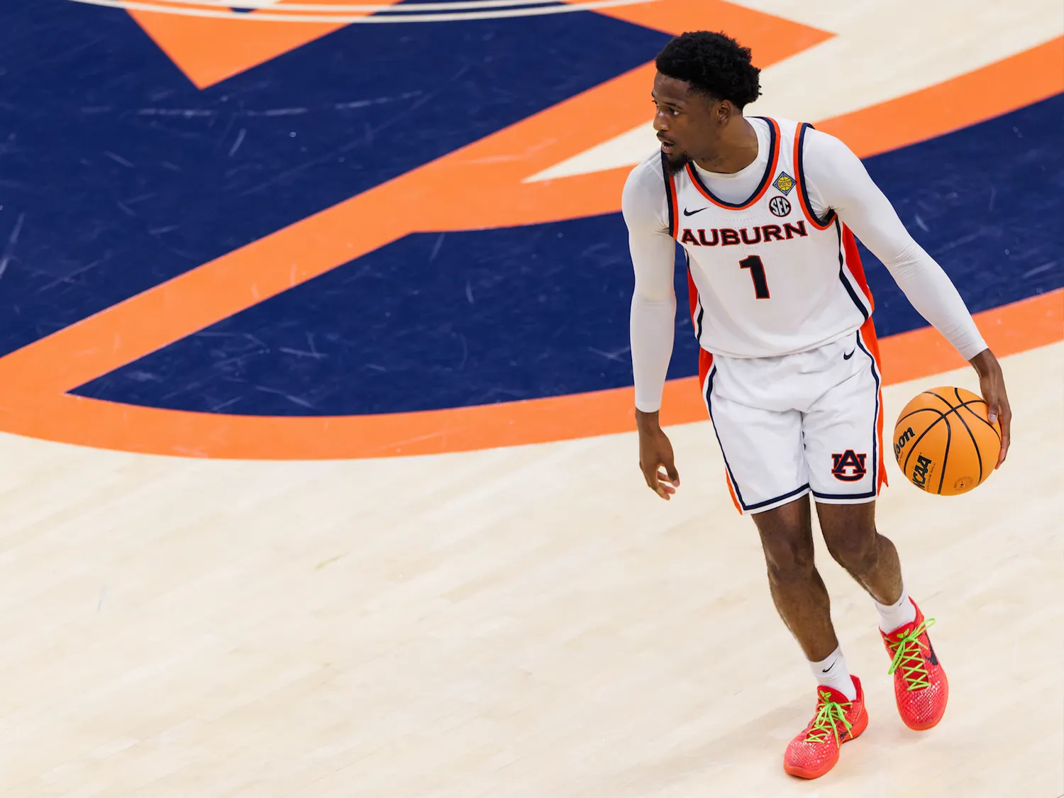 A male Auburn basketball player wearing a white jersey with the number 1 dribbles a ball on a hardwood court, looking to his left. He stands near a large orange and blue Auburn logo, wearing bright orange shoes with neon green laces.