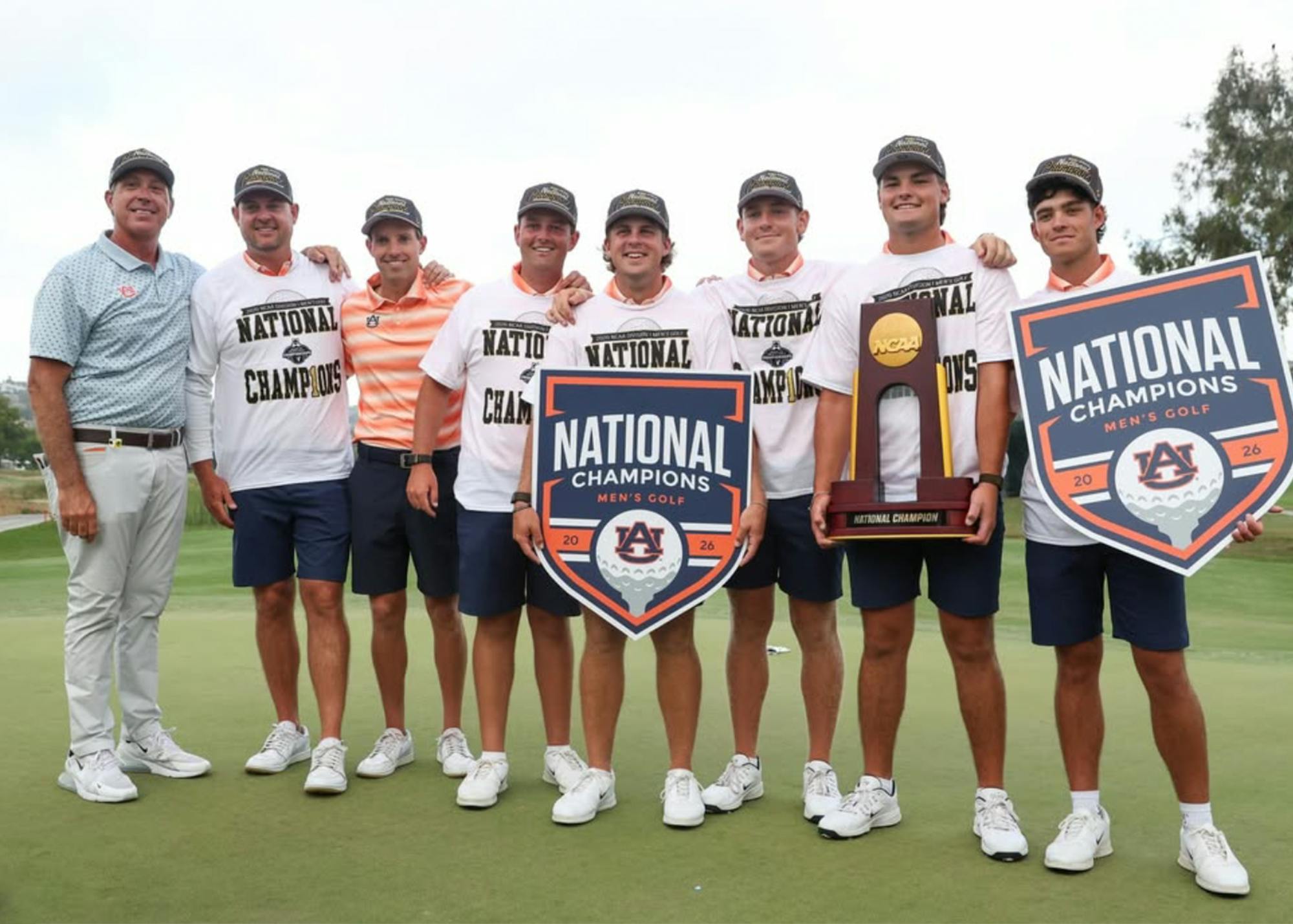 Auburn men’s golf players and coaches stand together on the green after winning the 2026 NCAA national championship. Several team members wear white “National Champions” shirts and championship hats while holding NCAA trophy hardware and large championship signs. The group smiles for a celebratory team photo following Auburn’s 4-1 victory over UCLA, securing the program’s second national title in three years and second national championship in school history.