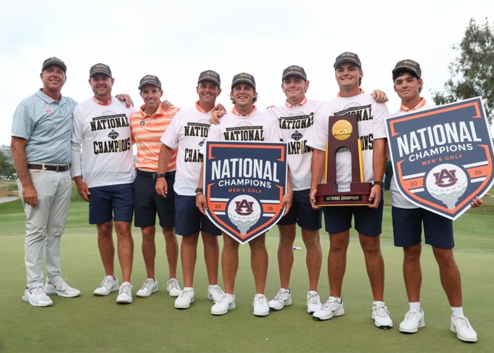 Auburn men’s golf players and coaches stand together on the green after winning the 2026 NCAA national championship. Several team members wear white “National Champions” shirts and championship hats while holding NCAA trophy hardware and large championship signs. The group smiles for a celebratory team photo following Auburn’s 4-1 victory over UCLA, securing the program’s second national title in three years and second national championship in school history.