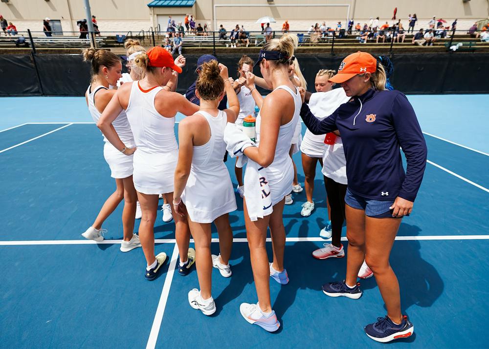 Auburn women’s tennis players gather in a huddle with a coach on the court during a break in play.