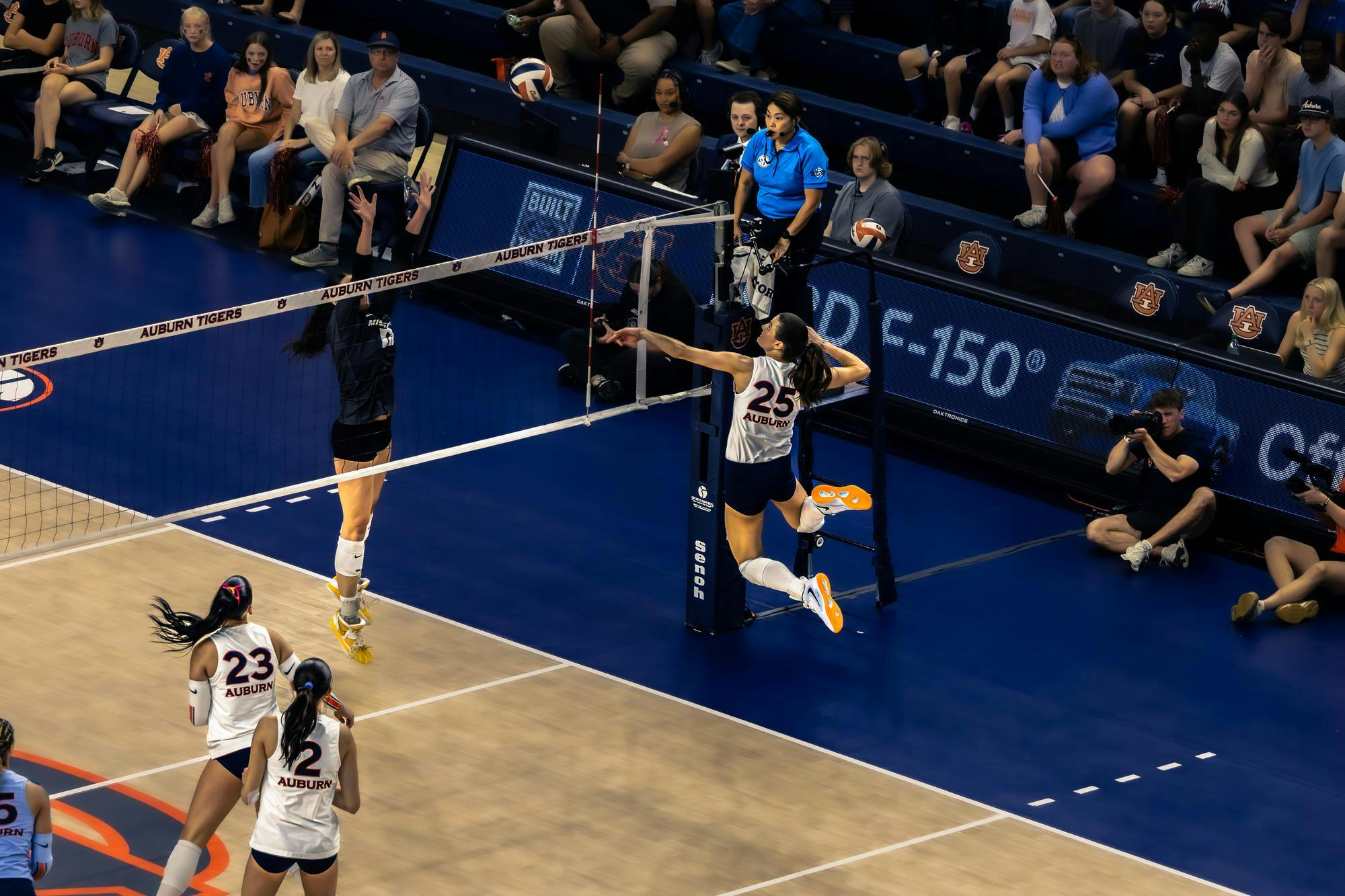 A volleyball player wearing a white Auburn jersey with number 25 jumps near the net to spike the ball as an opposing player in a dark uniform attempts to block. Teammates in white jerseys watch from the court, while spectators, photographers, and referees observe the play from the sidelines in an indoor arena with Auburn Tigers branding.