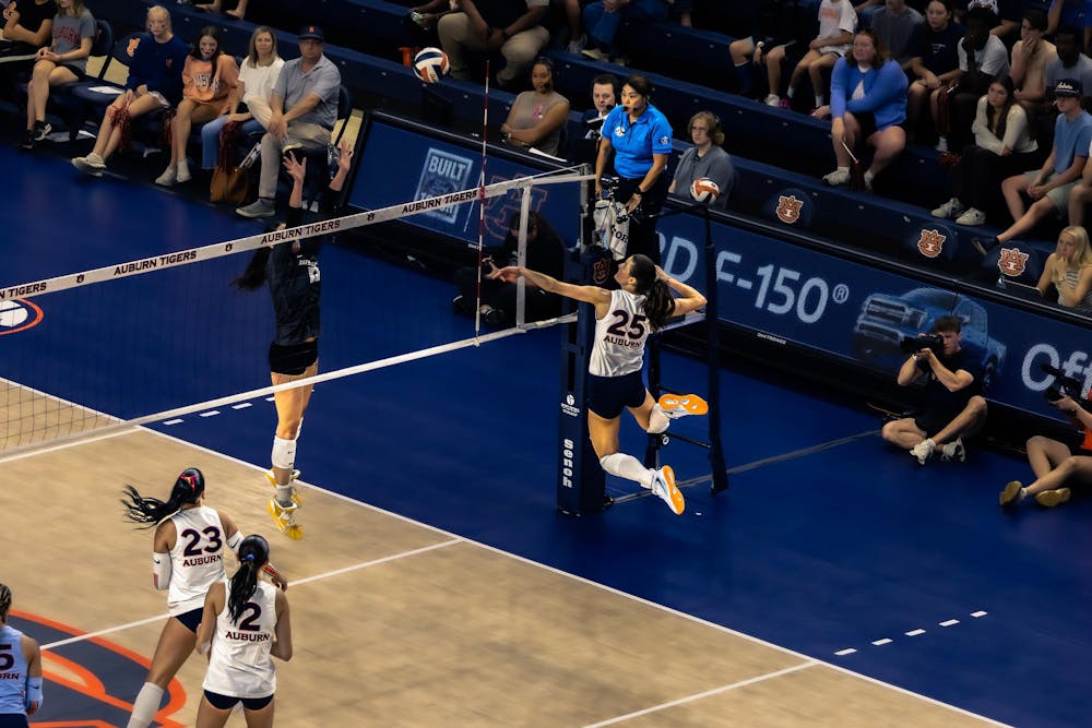 A volleyball player wearing a white Auburn jersey with number 25 jumps near the net to spike the ball as an opposing player in a dark uniform attempts to block. Teammates in white jerseys watch from the court, while spectators, photographers, and referees observe the play from the sidelines in an indoor arena with Auburn Tigers branding.