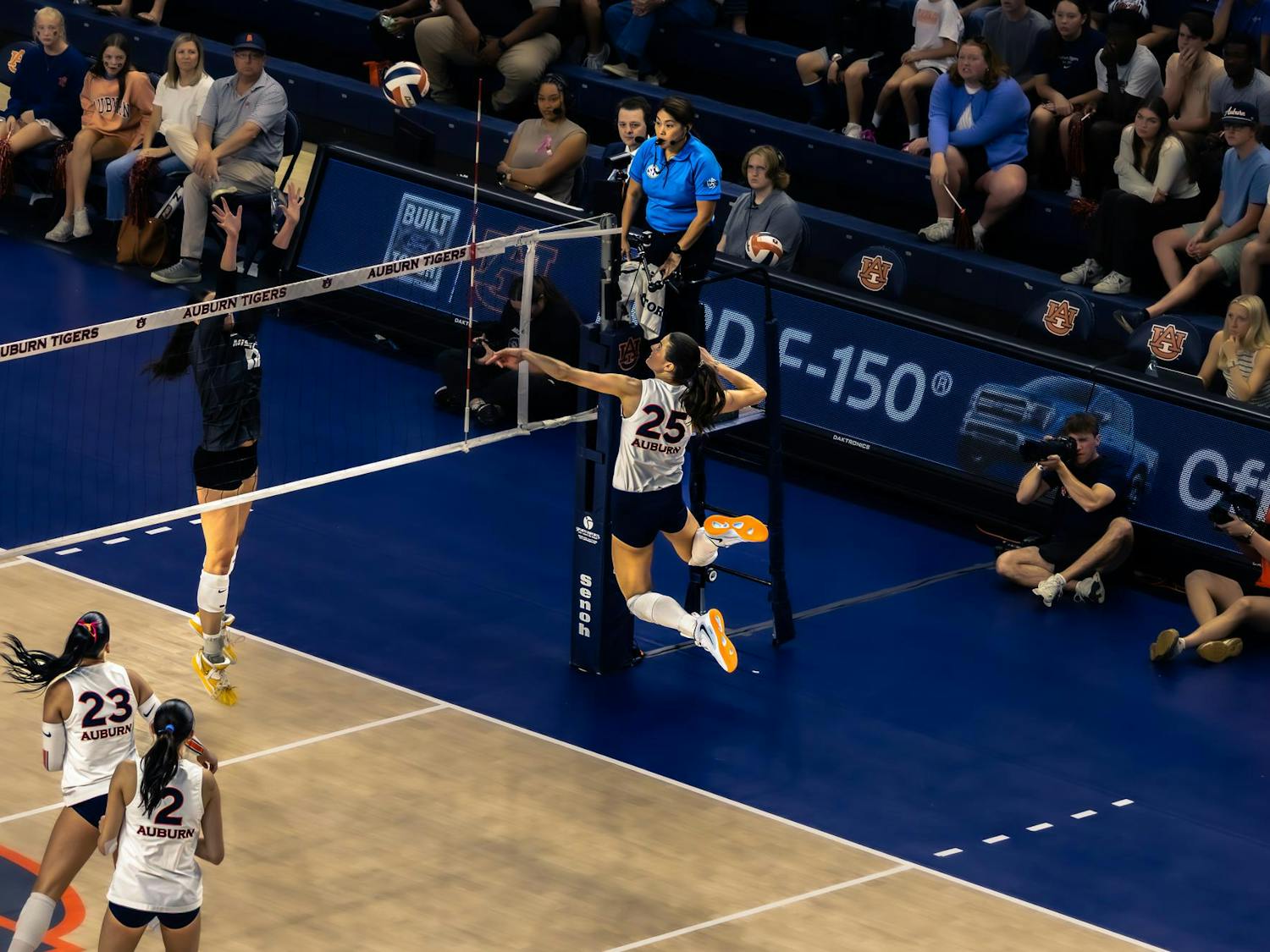 A volleyball player wearing a white Auburn jersey with number 25 jumps near the net to spike the ball as an opposing player in a dark uniform attempts to block. Teammates in white jerseys watch from the court, while spectators, photographers, and referees observe the play from the sidelines in an indoor arena with Auburn Tigers branding.