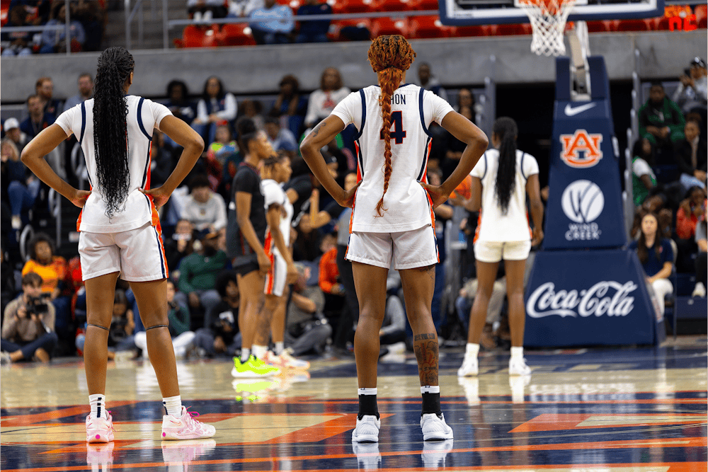 Four women’s basketball players in white Auburn uniforms stand on the court with their backs to the camera during a game. Two players in the foreground have their hands on their hips, with long braided hair visible. A basketball hoop, Auburn and Coca-Cola logos, and a crowd of spectators fill the background.