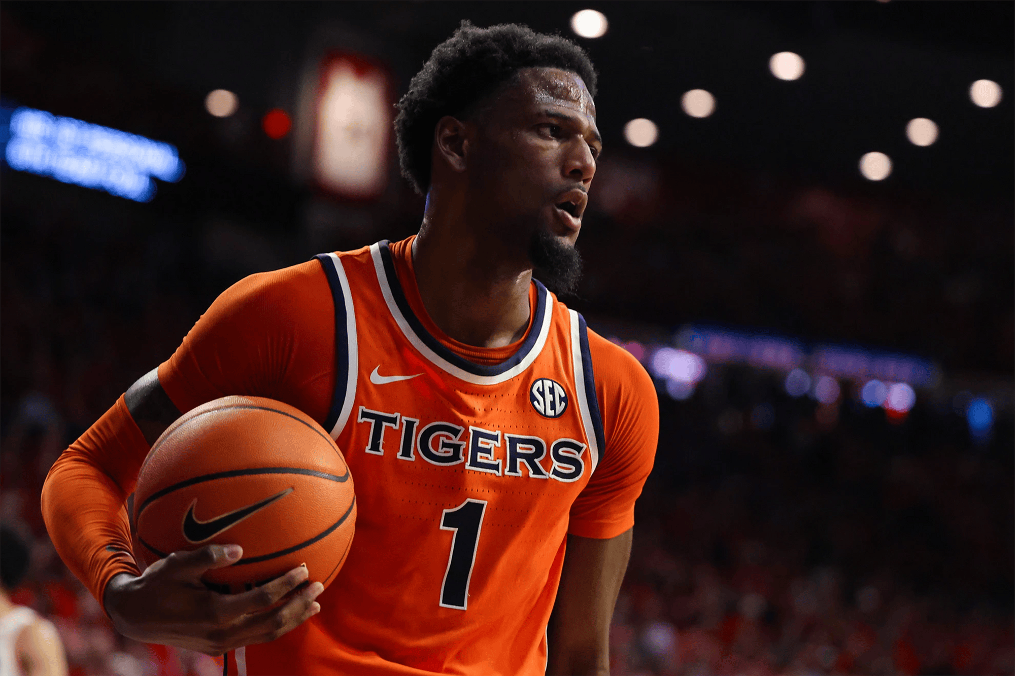 Auburn men’s basketball player in an orange No. 1 “Tigers” jersey holds the ball at his hip while looking across the court, with arena lights and a blurred crowd in the background.