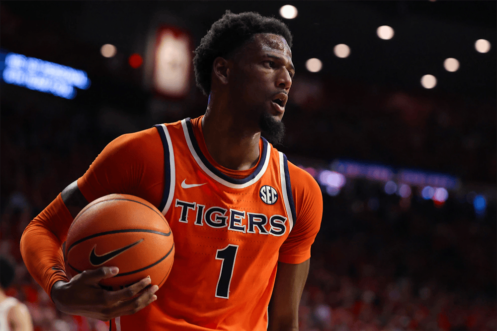 Auburn men’s basketball player in an orange No. 1 “Tigers” jersey holds the ball at his hip while looking across the court, with arena lights and a blurred crowd in the background.