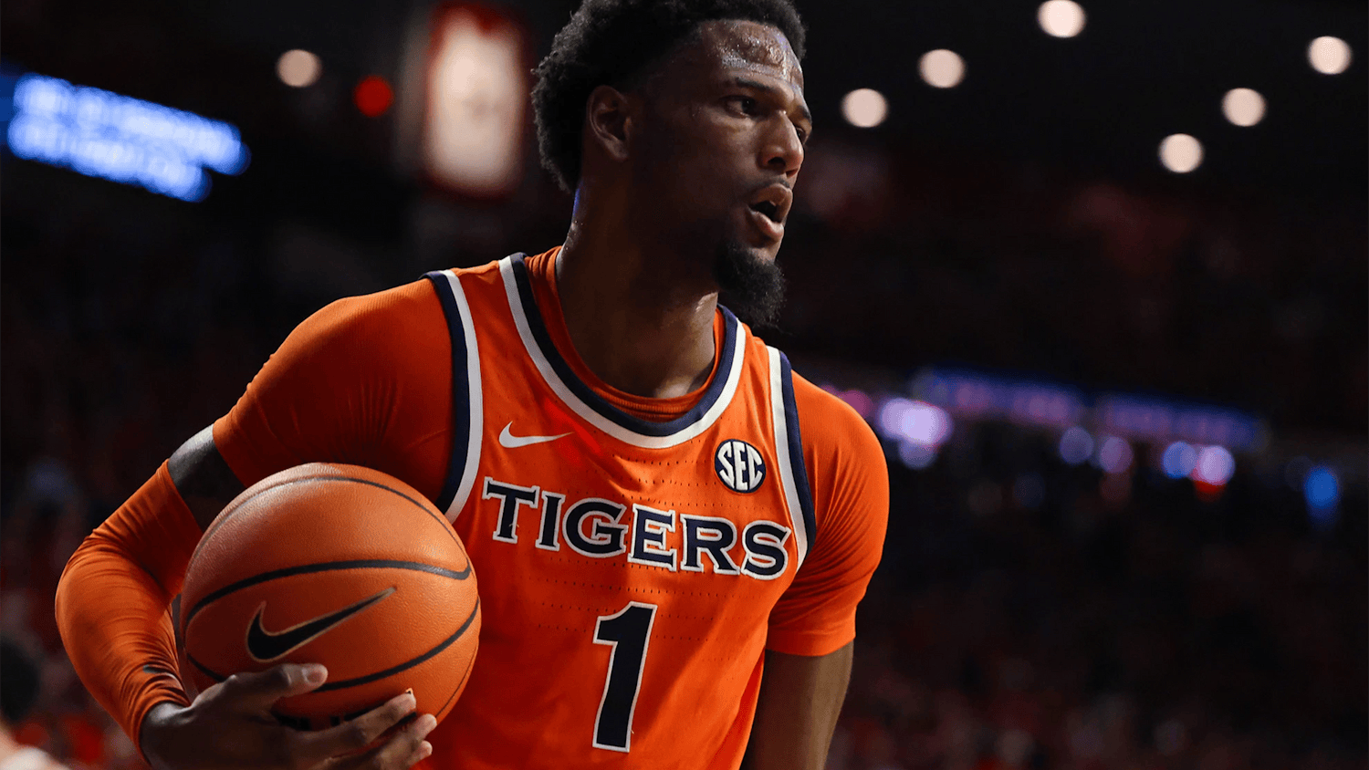 Auburn men’s basketball player in an orange No. 1 “Tigers” jersey holds the ball at his hip while looking across the court, with arena lights and a blurred crowd in the background.