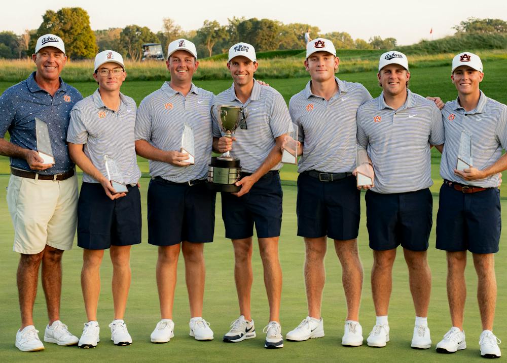 Auburn men's golf coach and six players pose on a golf course holding trophies after winning the Inverness Intercollegiate.