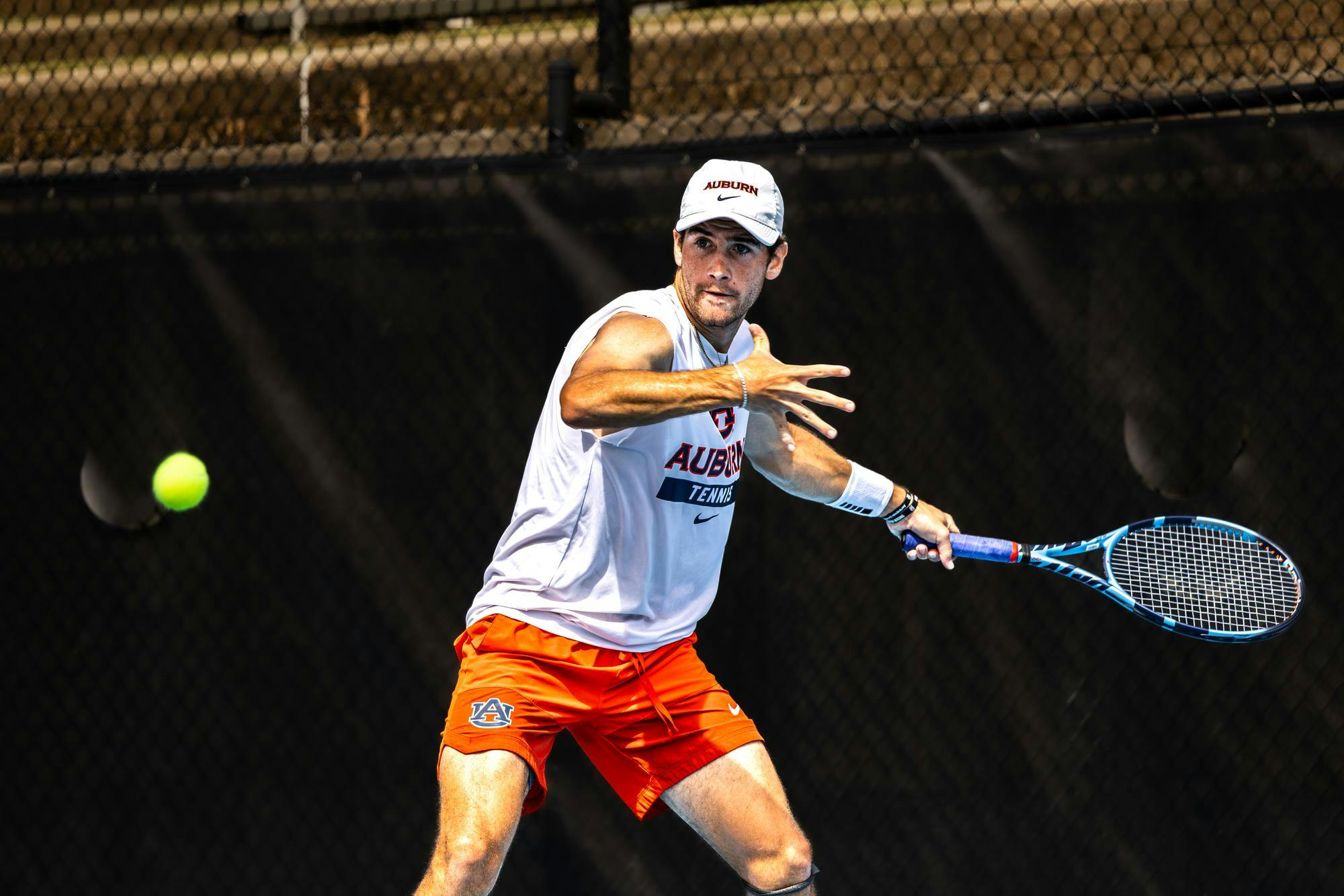 Auburn men’s tennis player prepares to hit a forehand shot during a match, wearing an Auburn cap, white sleeveless shirt, and orange shorts, with a focused expression on an outdoor court.