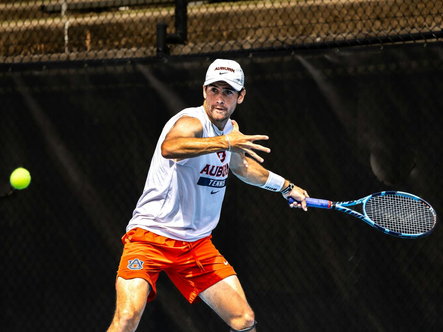 Auburn men’s tennis player prepares to hit a forehand shot during a match, wearing an Auburn cap, white sleeveless shirt, and orange shorts, with a focused expression on an outdoor court.