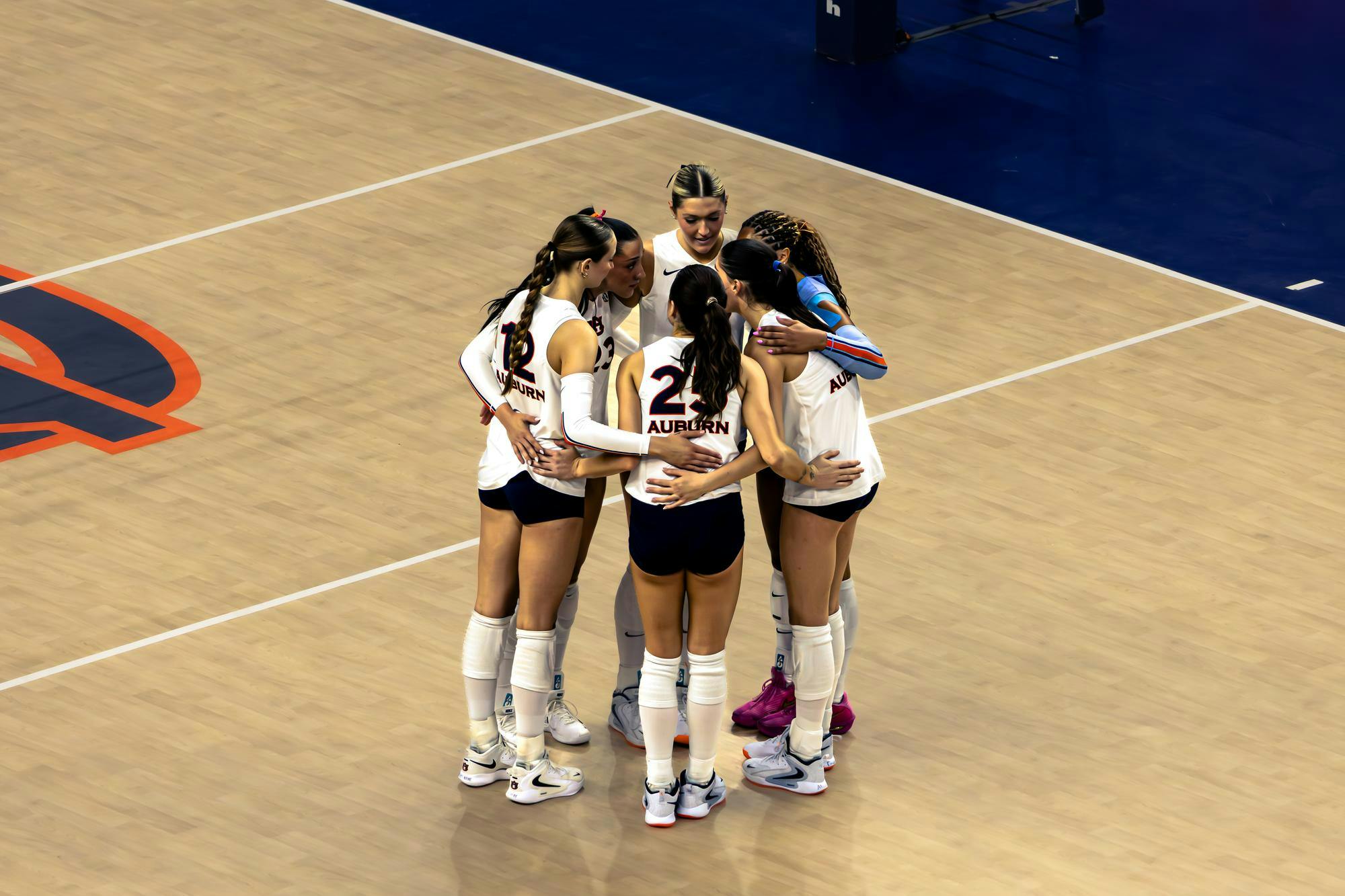 A group of six Auburn volleyball players wearing white jerseys and navy shorts huddle together on the court, arms around each other, before the start of a play.