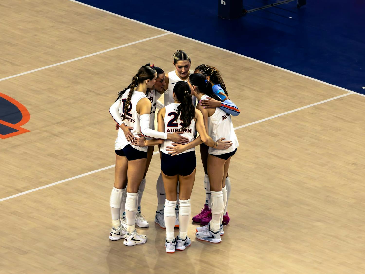 A group of six Auburn volleyball players wearing white jerseys and navy shorts huddle together on the court, arms around each other, before the start of a play.