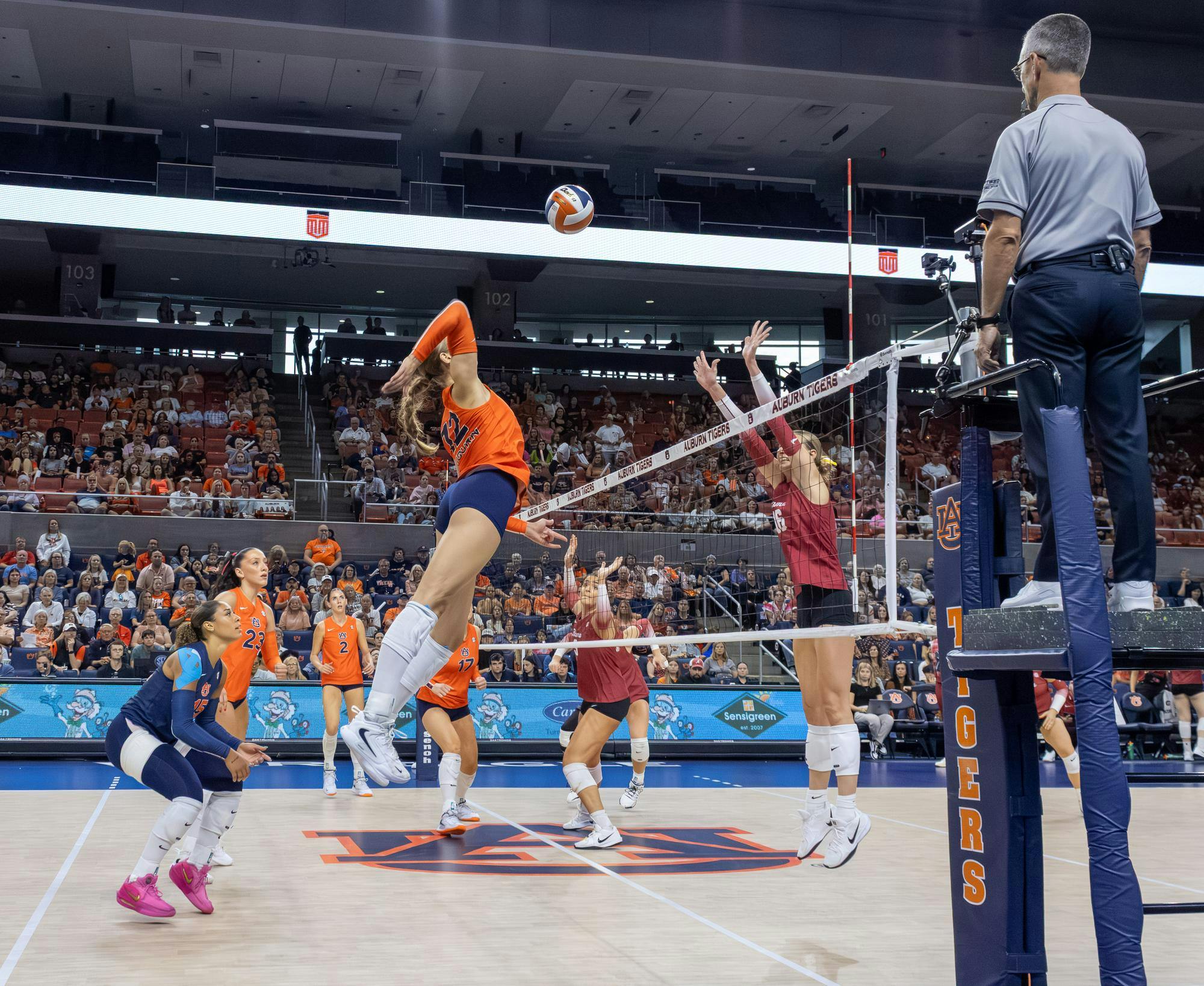 Auburn volleyball player in an orange jersey jumps high for a spike against Alabama defenders at the net, as teammates and the referee look on in a packed arena.
