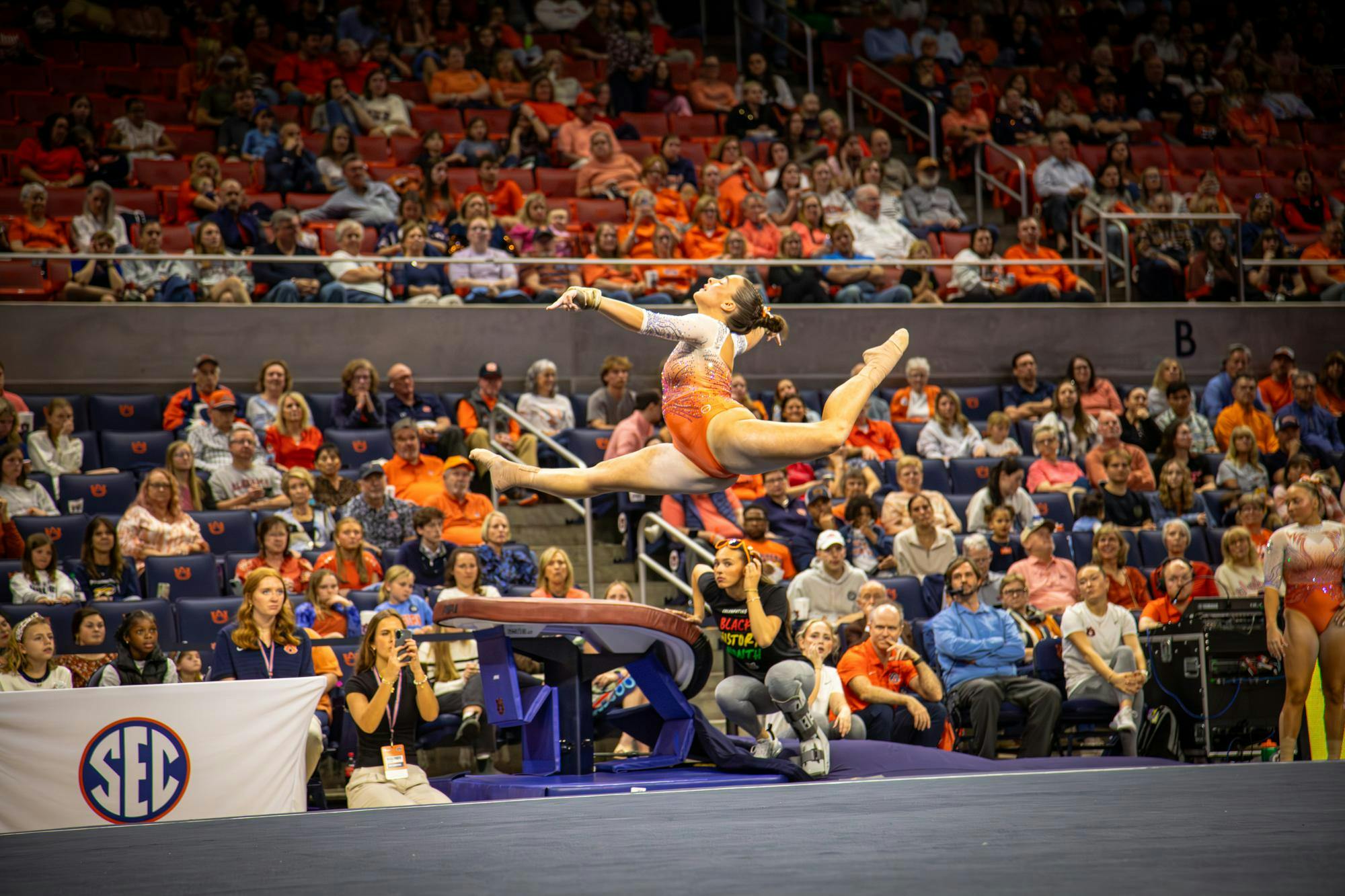 Auburn gymnast in an orange leotard performs a split leap during a floor routine in front of a large crowd, with judges, staff, and SEC signage visible courtside.