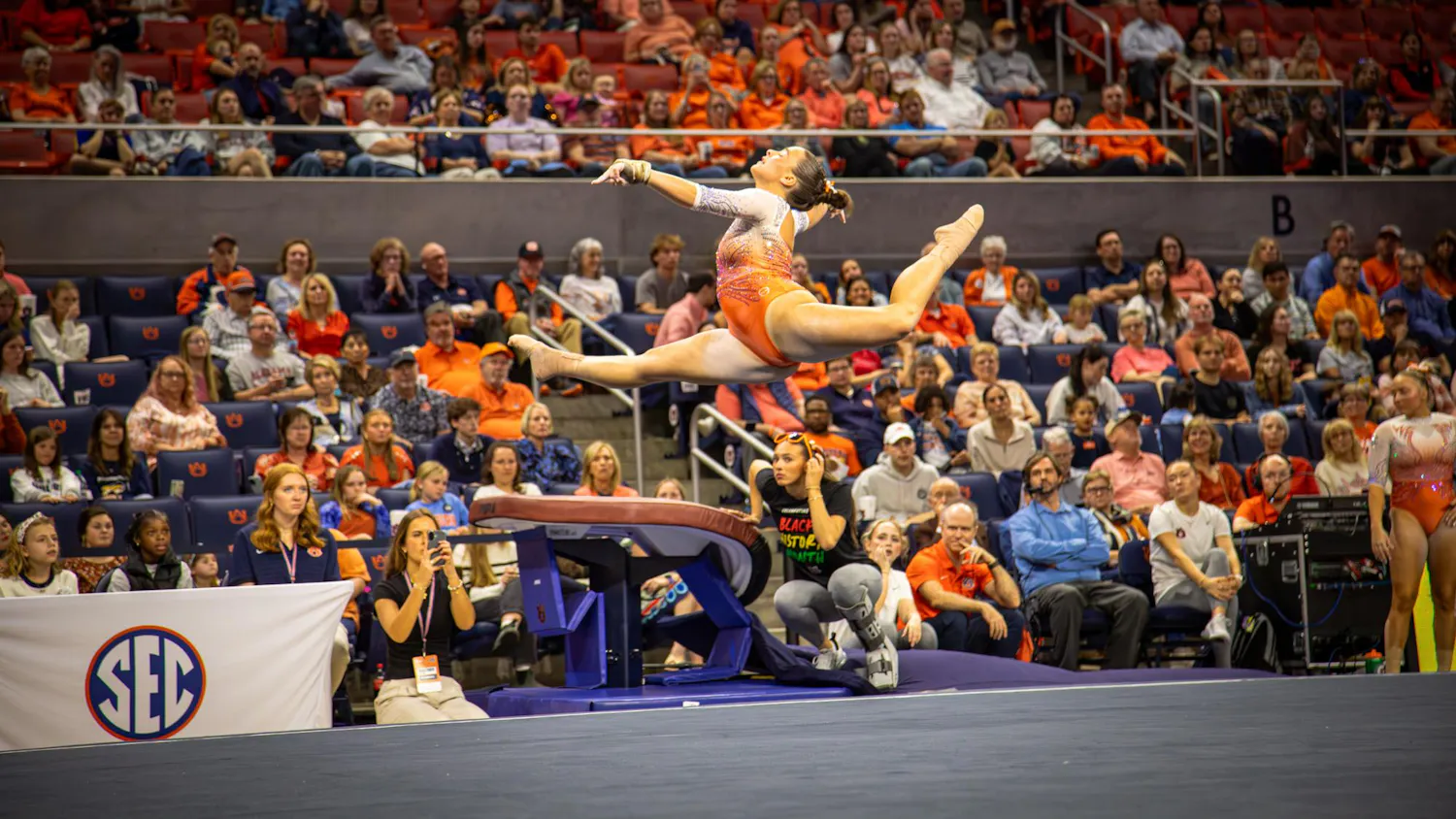 Auburn gymnast in an orange leotard performs a split leap during a floor routine in front of a large crowd, with judges, staff, and SEC signage visible courtside.
