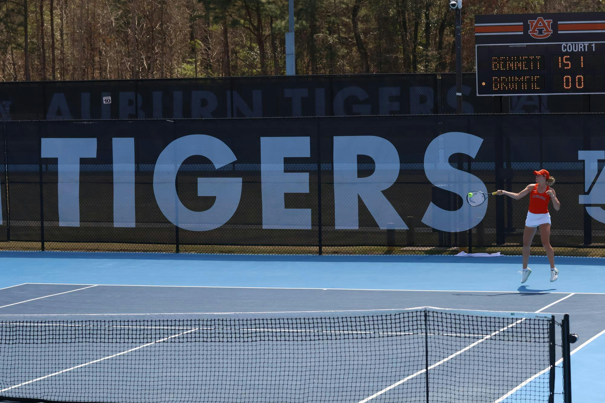 Auburn's DJ Bennett serves on a blue outdoor court while standing under a scoreboard.